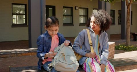 Asian and african american teenage students sitting on campus bench searching backpack