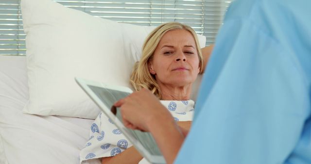 A middle-aged Caucasian woman in a hospital gown is looking at a digital tablet, with copy space. She appears to be a patient receiving information or entertainment during her healthcare stay.