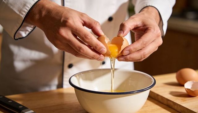 Chef cracking egg into enamel bowl on rustic wooden countertop close-up