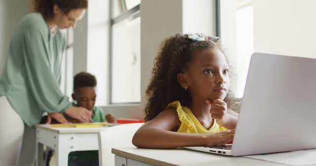 An African American girl in a yellow dress using a laptop in a brightly lit classroom. She appears thoughtful, resting her chin on her hand. A teacher in green cardigan assists another student in focus. This image is perfect for illustrating concepts of modern education, digital learning, and classroom dynamics. Useful for educational websites, learning materials, and advertisements related to children's education.