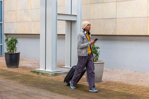 Young asian traveler walking pulling suitcase while holding smartphone near modern building