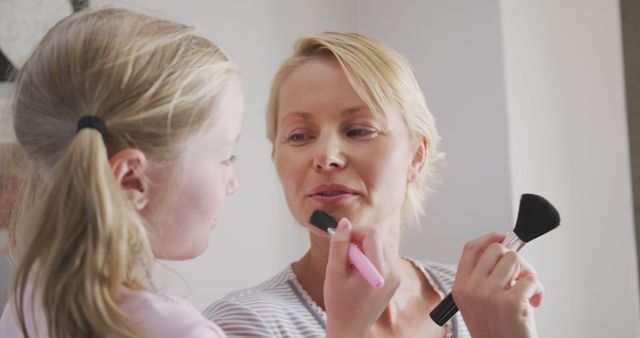 Happy caucasian daughter having fun using makeup brush on face of her smiling mother. Motherhood, childhood, domestic life, care, fun and togetherness.