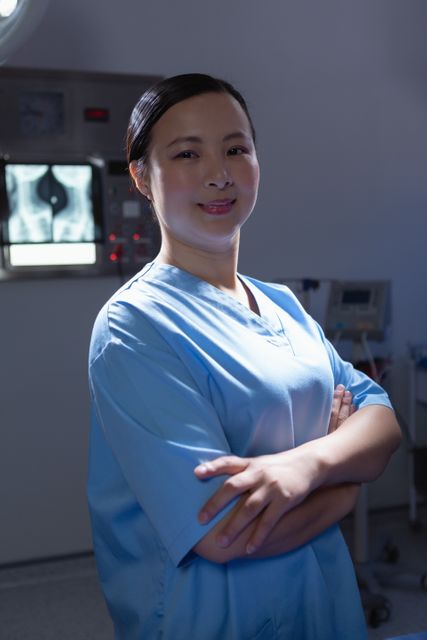 A confident female surgeon stands with arms crossed, looking directly at the camera in an operating room. She is wearing blue scrubs, and medical equipment is visible in the background. This image can be used for healthcare-related content, medical websites, hospital brochures, and articles about medical professionals and surgery.