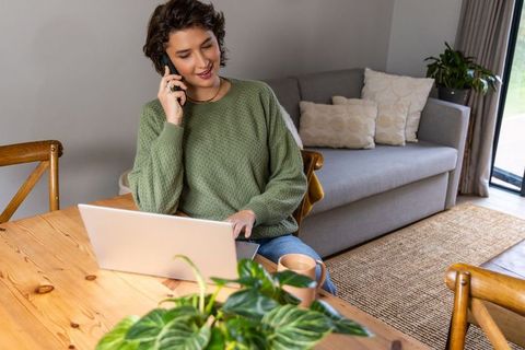 Woman working from home, multitasking on laptop and phone at cozy living room table