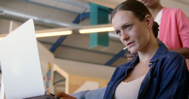 Young woman concentrating on her laptop in a modern office, with casual clothing suggesting a relaxed workplace. Ideal for articles on productivity, women in technology, or modern work environments.