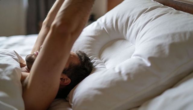 Shirtless man lying in bed stretching arms overhead on large supportive pillow and cozy duvet