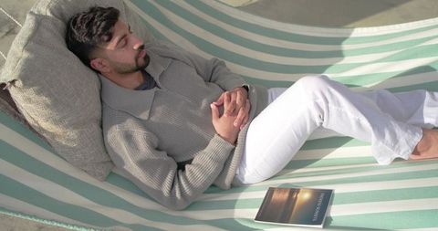 Indian man reclining in striped hammock on sunlit terrace wearing sweater and white pants