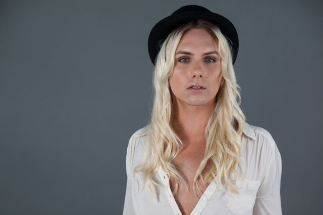 Transgender woman with blonde hair wearing a black hat and white shirt, standing against a gray background. Ideal for use in articles about gender identity, fashion, individuality, and confidence. Suitable for websites, magazines, and social media posts focusing on LGBTQ+ topics and personal expression.
