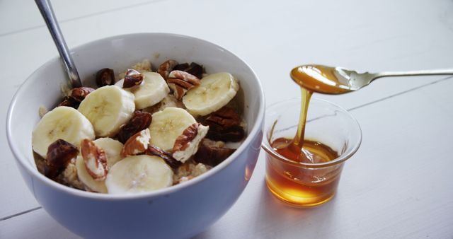 A bowl of oatmeal topped with banana slices and pecans sits next to a small dish of honey, with copy space. Honey is being drizzled from a spoon, adding a sweet touch to this nutritious breakfast setup.