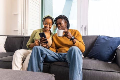 African american couple relaxing on sofa, sharing smartphone and coffee in sunlit living room