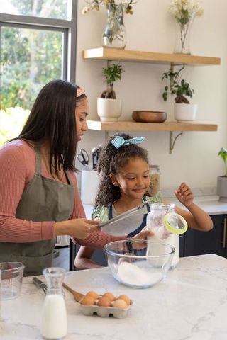 African american mother and daughter baking together in sunlit kitchen, sifting flour