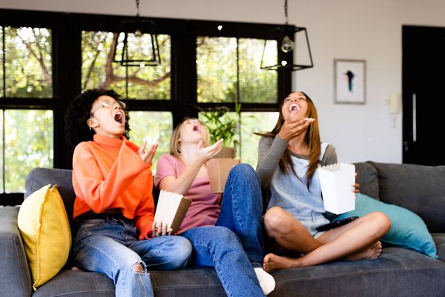 Three teenage girls of diverse backgrounds are sitting on a couch, enjoying popcorn and watching TV. They are laughing and having fun, creating a lively and joyful atmosphere. This image can be used for promoting home entertainment, friendship, and leisure activities. It is ideal for advertisements, social media posts, and articles related to teenage life, bonding, and relaxation.