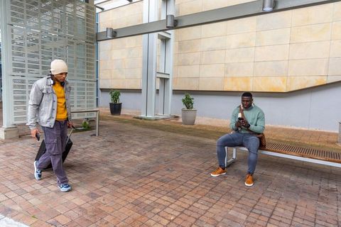 Young diverse men commuting in urban plaza using smartphones and pulling rolling luggage