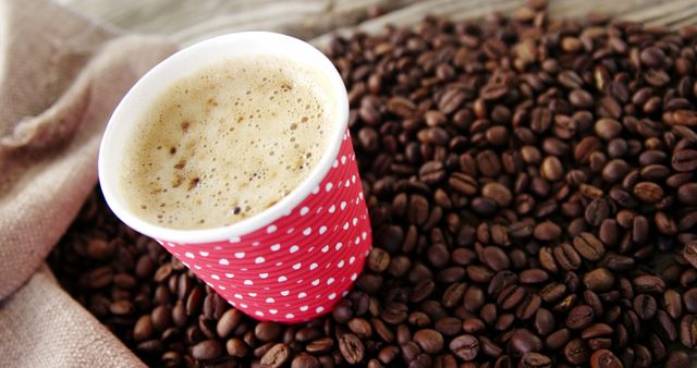 A red polka-dotted paper cup filled with frothy coffee sits atop a bed of roasted coffee beans, with copy space. The image evokes the aroma and warmth of a freshly brewed cup of coffee, perfect for coffee enthusiasts.