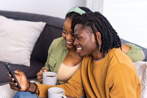 African american couple sharing coffee and laughing while looking at smartphone on sofa