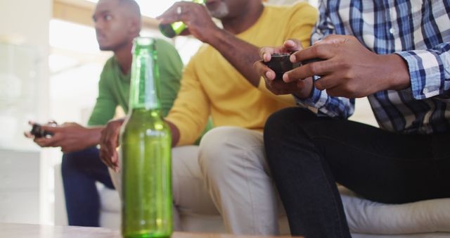 Three men are sitting on a couch in a living room, playing video games and drinking beer. The foreground features a bottle of beer on a table, suggesting a relaxed, casual environment. Perfect for themes of friendship, leisure, bonding, and modern relaxation.