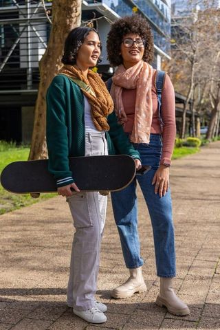 Urban skateboard friends holding board and laughing on sunlit campus wearing scarves and denim