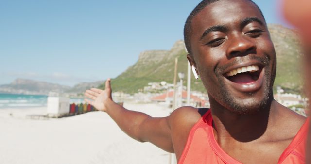 A young man in a red tank top taking a selfie on a sunny beach with colorful huts in the background. He appears happy and relaxed, with mountains and ocean adding to the scenic view. This image is perfect for travel promotions, vacation brochures, social media content related to joy and leisure, or campaigns focusing on outdoor activities and beach holidays.