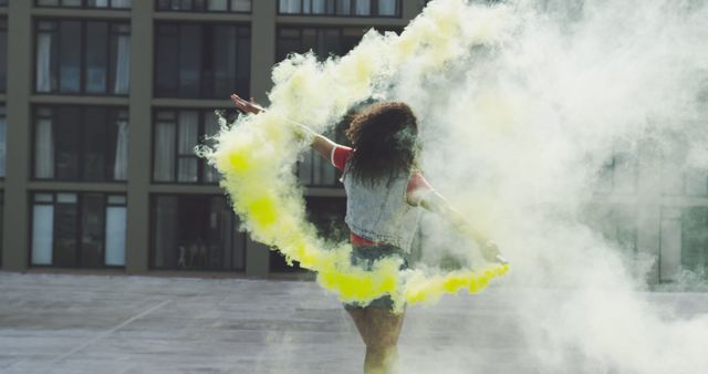 Young biracial woman dances amidst vibrant yellow smoke outdoors. Her dynamic movement creates an artistic contrast with the urban backdrop.