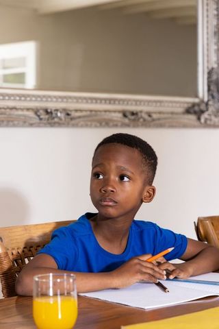 African american boy doing homework at wooden dining table holding pencil and notebook