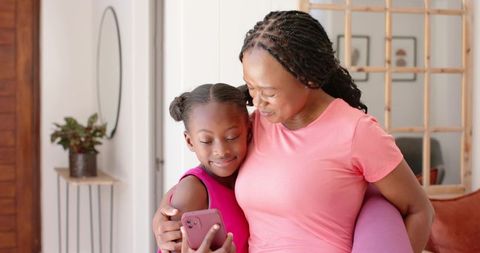 African american mother hugging daughter holding pink phone and yoga mat, sharing smile