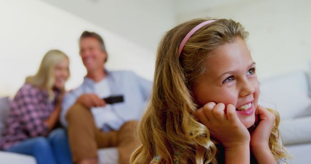 Young girl with blonde hair and headband smiles while watching TV. In the blurred background, her parents sit relaxed on the sofa, enjoying family time. The setting depicts a warm and happy moment at home, ideal for family life, togetherness, and living room scenes. Perfect for uses in advertisements, blogs on family bonding, children's activities, and home interior concepts.