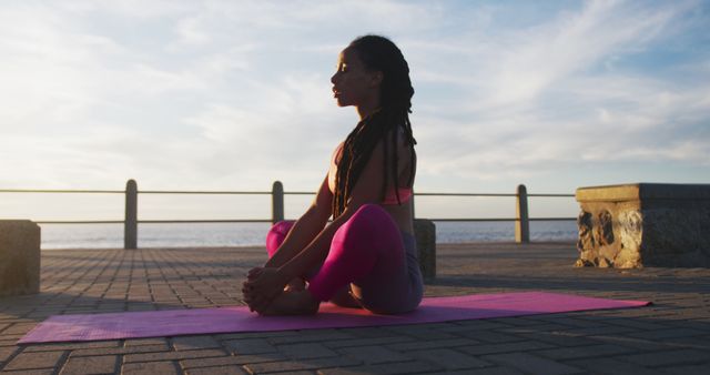 Female yoga practitioner sitting in a meditation pose on a pink yoga mat near the seaside, likely at sunrise. Ideal for promoting wellness, fitness, mindfulness, and outdoor exercise. Suitable for use in health and fitness blogs, mental well-being articles, yoga class advertisements, or meditation guides.