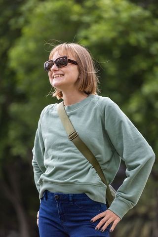 Young woman smiling and wearing sunglasses, posing hands-on-hips in green park