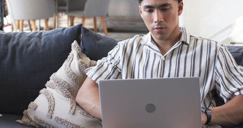 Asian man working from home on laptop while lounging on gray sofa with textured pillow