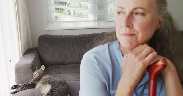 Senior woman sitting on couch, holding a cane, looking pensive. A grey tabby cat sleeps on the couch next to her. Using this image emphasizes themes of companionship, relaxation, and domestic life. Useful for illustrating topics related to aging, pets, home care, or peaceful moments indoors.