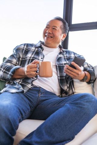 Mature Asian Man Relaxing Holding Coffee Mug And Smartphone