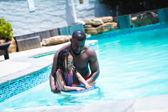 African American father helping his young daughter learn to swim in a pool on a sunny day. Ideal for use in content related to family bonding, parenting, summer activities, outdoor fun, and lifestyle. Perfect for advertisements, blogs, and articles focusing on family life, swimming lessons, and leisure activities.