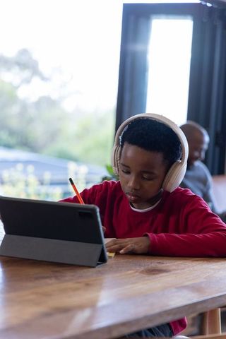 African american boy studying with tablet and headphones doing homework by sunlit window