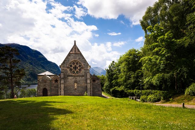 This photo features a historic stone church set against a backdrop of lush greenery and scenic mountains in daytime. The clear sky and verdant landscape create a serene and majestic atmosphere, making this perfect for concepts related to heritage, spirituality, and peaceful retreats. Suitable for use in travel brochures, spiritual websites, historical documentation, and countryside tourism promotions.