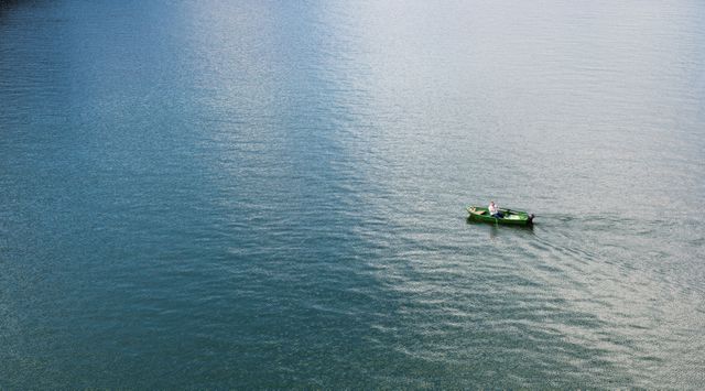 This image shows a lone fisherman rowing a small green boat on calm open water, suitable for illustrating themes of solitude, peace, and nature's serenity. Great for websites, blogs, and articles related to fishing, nature, tranquility, and personal reflection. Ideal for use in travel brochures and outdoor adventure promotions.