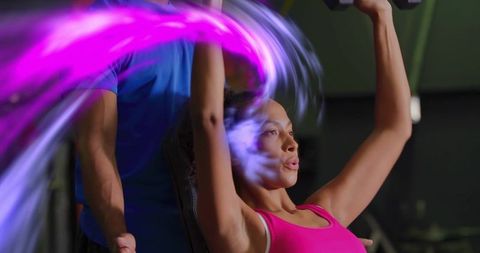 Female athlete performing dumbbell overhead press with trainer, neon motion trail