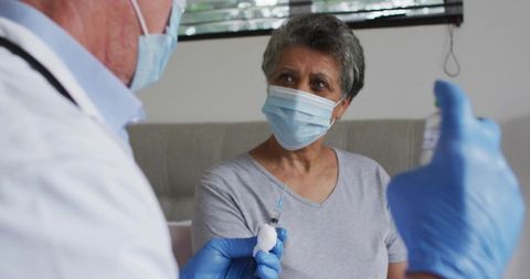 Mature woman receiving vaccine while masked doctor preparing syringe and vial in clinical care