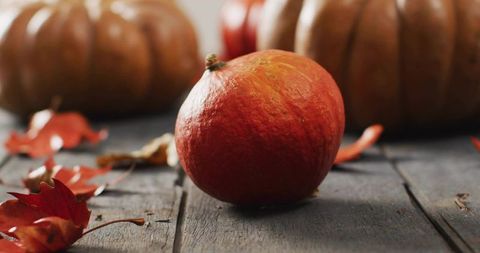 Small orange pumpkin sitting on rustic wooden table with autumn leaves and blurred gourds