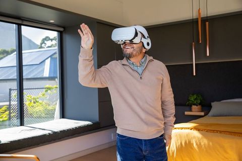 Mature asian man wearing vr headset exploring virtual reality in modern sunlit bedroom