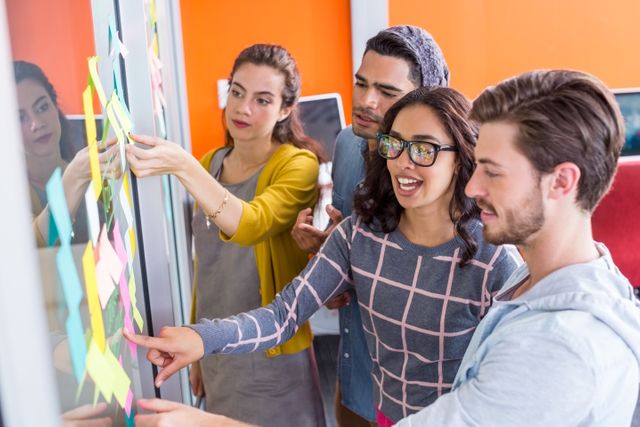 Group of young professionals collaborating and brainstorming using sticky notes on a glass wall. Ideal for illustrating teamwork, creativity, and collaborative work environments in modern offices. Can be used in business presentations, team-building articles, or workplace strategy guides.
