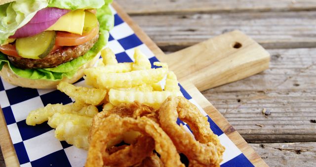 A delicious-looking burger with lettuce, tomato, and cheese is served alongside crispy fries and onion rings on a wooden board with a checkered paper. The meal presents a classic American fast food experience, tempting with its combination of textures and flavors.