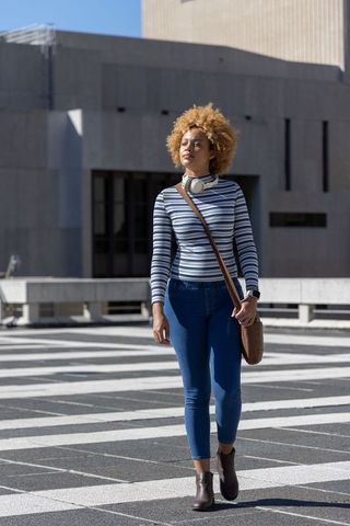 African american woman walking across urban plaza with headphones and brown crossbody bag