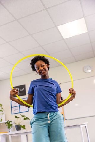 Joyful school-age boy holding bright yellow hula hoop in classroom, smiling brightly