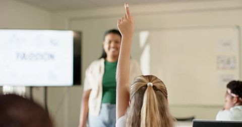 Student raising hand in classroom while teacher leads lesson with laptop display