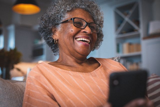 Elderly African American woman sitting in living room, smiling while using smartphone. Ideal for content related to senior lifestyle, technology use among elderly, happiness, home comfort, and family communication.