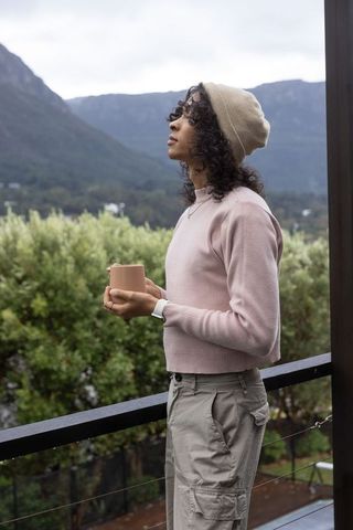 Non-binary person standing on balcony holding mug and gazing at misty mountain valley
