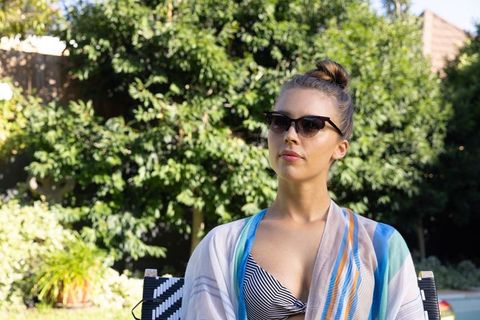Chic woman relaxing on patio in backyard wearing cat-eye sunglasses and striped bikini
