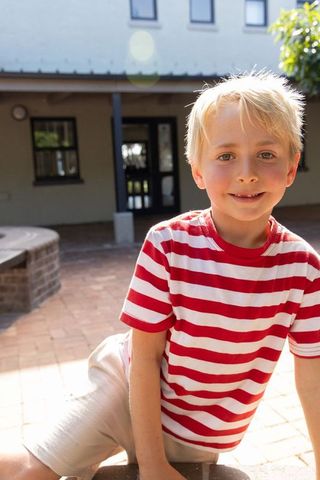 Sunlit blonde boy leaning on brick planter in courtyard wearing red striped tee, smiling