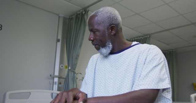 Elderly man with a beard standing next to a hospital bed in a gown, looking observed. Can be used in health and medicine projects, illustrating patient care, hospital environments, and patient recovery imagery.