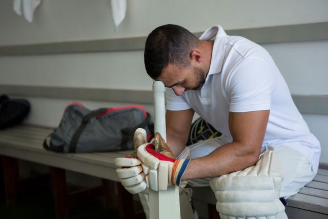This image depicts a cricket player sitting on a bench in a locker room, appearing stressed and contemplative. He is dressed in full cricket gear, including gloves and pads, with a cricket bat resting between his legs. This image can be used in articles or advertisements related to sports psychology, athlete mental health, cricket training, or the pressures of competitive sports.